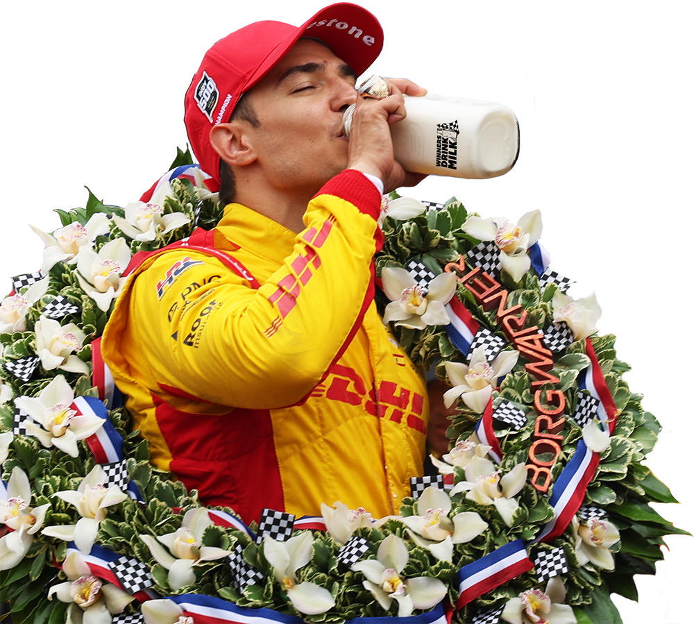 Indy 500 race winner Álex Palou in a yellow and red racing suit wearing a large floral wreath and red cap, tilting back a glass bottle of milk in Victory Lane against a white background.