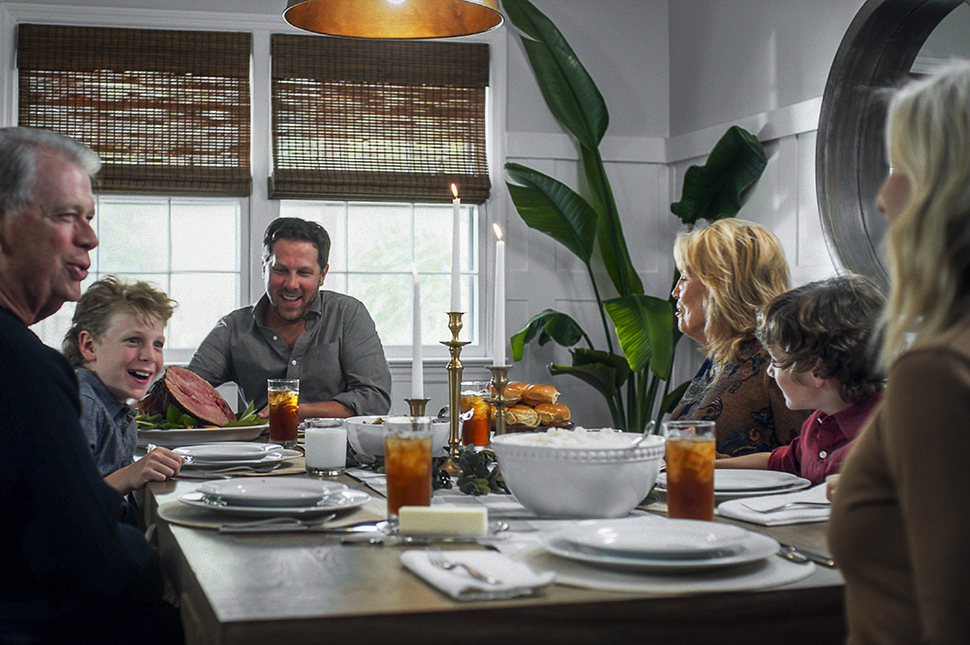 A family gathered around a warmly lit dining table, laughing and enjoying each other’s company as they prepare for a holiday meal. The table is set with plates, glasses of iced tea, a centerpiece with candles, and a large ham as the focal point, creating a cozy and inviting atmosphere.