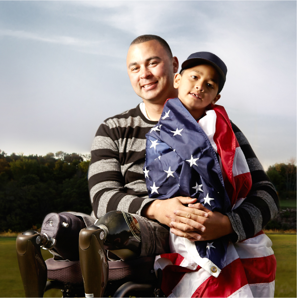 An adult with prosthetic legs sits outdoors holding a young child wrapped in an American flag, both looking toward the camera.
