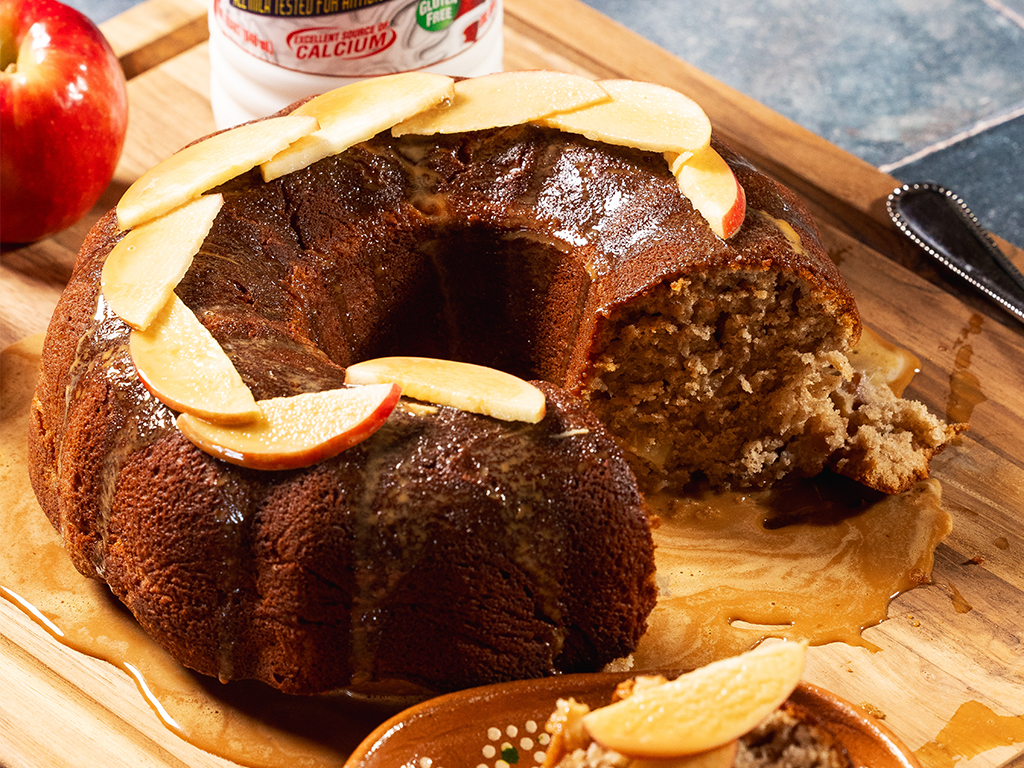 A sliced bundt apple cake topped with apple slices and drizzled with caramel glaze, displayed on a wooden cutting board, with an apple and a Hiland Dairy milk container in the background.