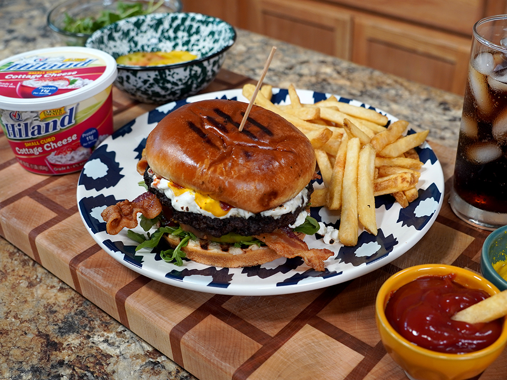 A loaded bacon cheeseburger with cottage cheese and a fried egg is served on a patterned plate alongside a generous portion of golden French fries, with a glass of iced cola, ketchup, mustard, and a tub of Hiland Cottage Cheese nearby on a wooden kitchen counter.