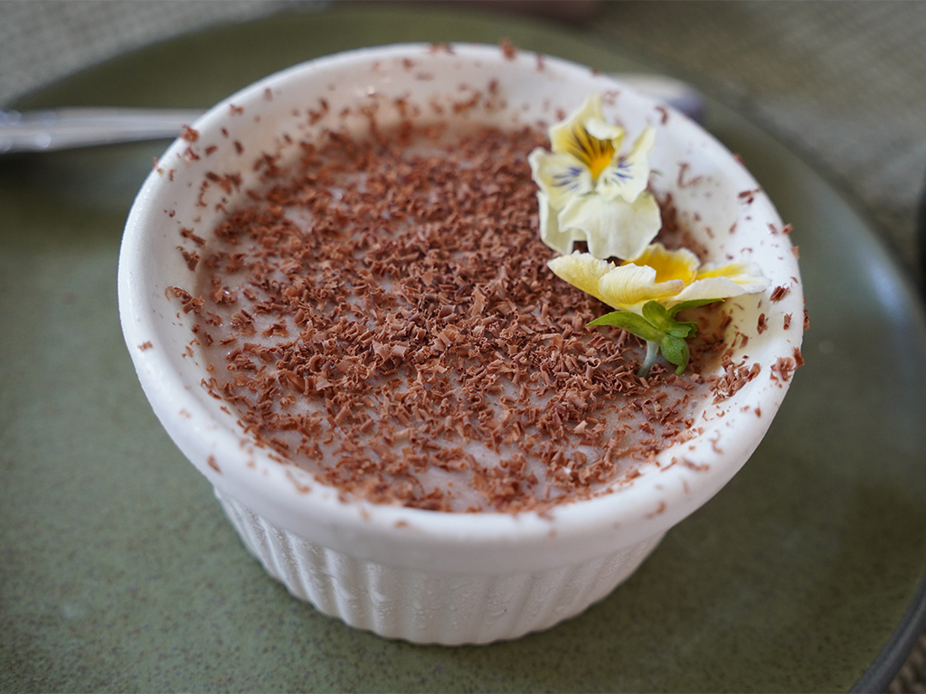 Chocolate pudding served in a white ramekin and topped with finely grated chocolate and edible flowers, presented on a green plate with a spoon in the background.