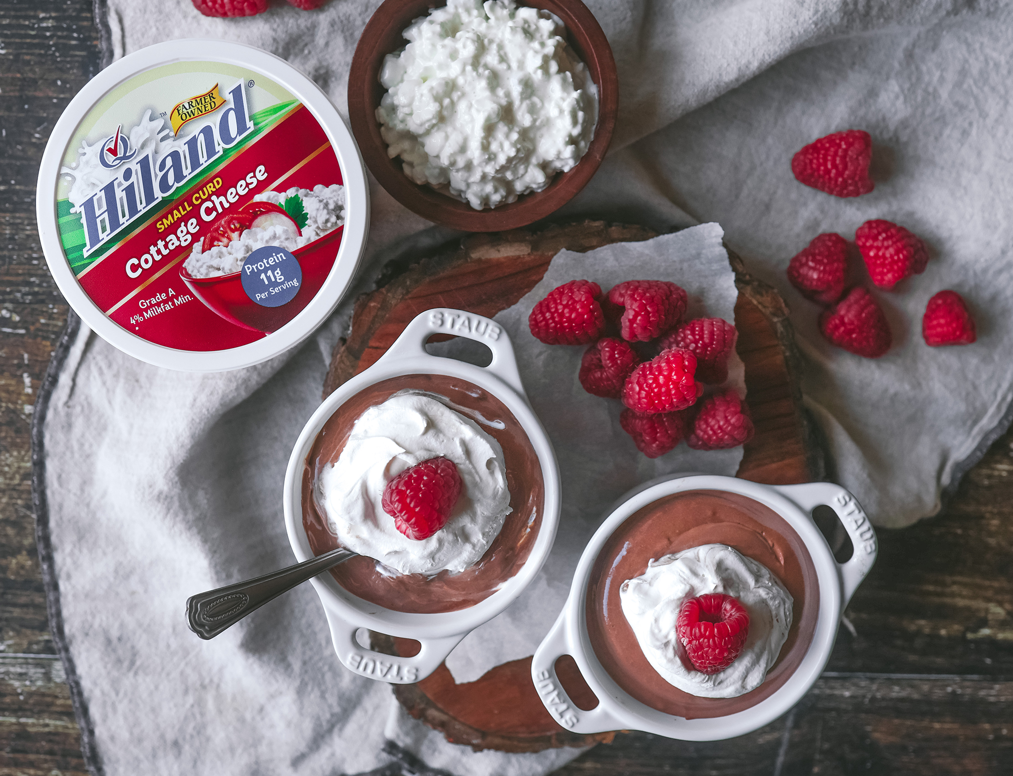 Cottage cheese chocolate mousse in small white bowls, displayed with a container of Hiland Cottage Cheese and fresh raspberries on a wooden backdrop.