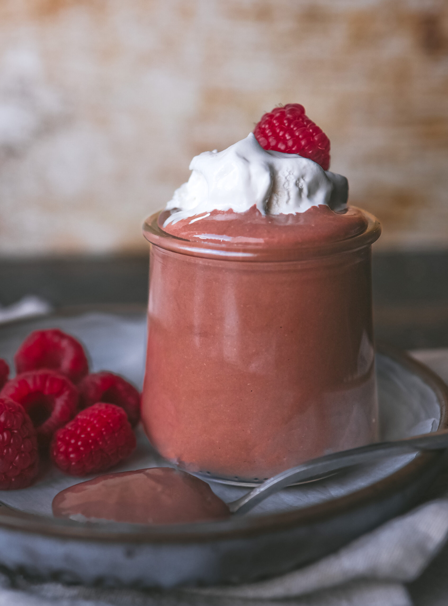 A single serving of chocolate mousse in a glass jar, topped with whipped cream and a raspberry, placed on a plate with scattered raspberries.