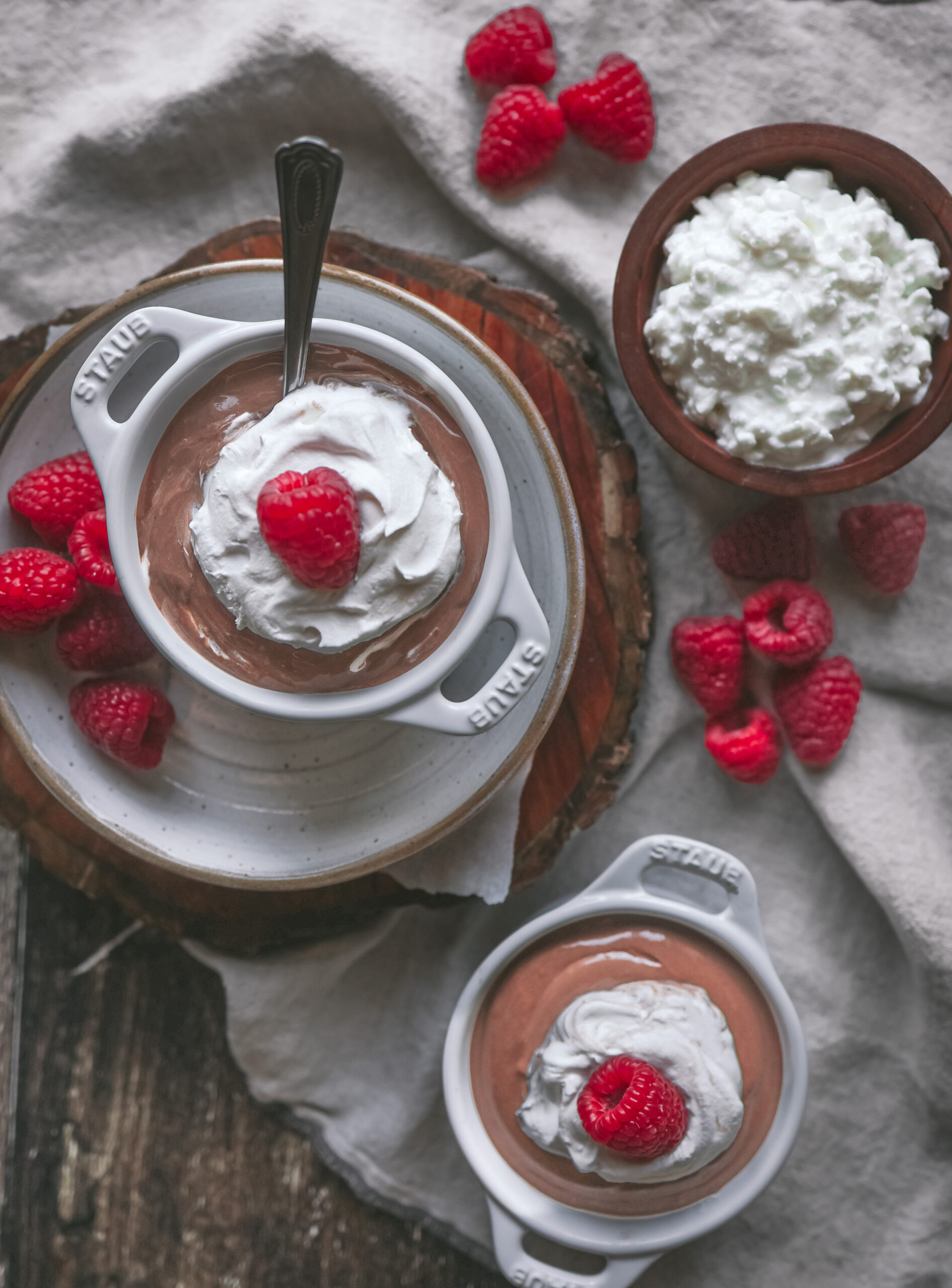 Overhead view of two bowls of chocolate mousse garnished with whipped cream and raspberries, accompanied by a wooden bowl of cottage cheese and fresh raspberries.