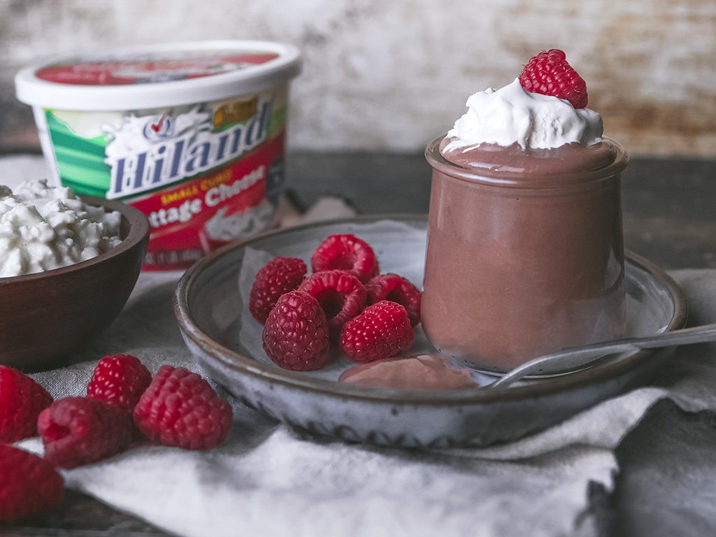 Chocolate mousse served in a glass jar with whipped cream and raspberries, accompanied by Hiland Cottage Cheese and fresh raspberries in a rustic setting.