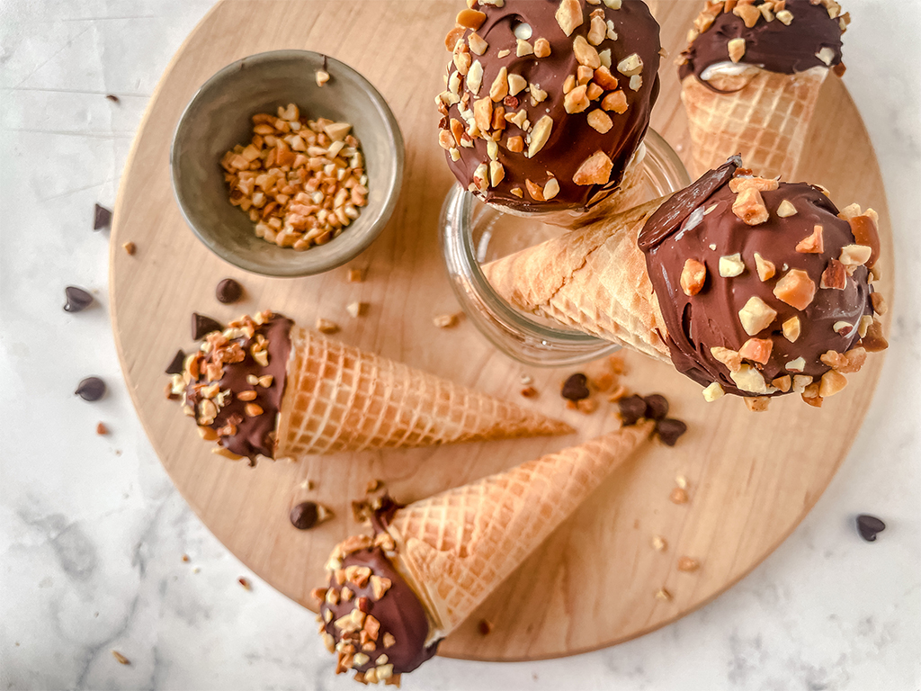 Homemade drumstick ice cream cones with chocolate-dipped tops coated in crushed nuts, displayed on a wooden board with extra nuts and chocolate chips scattered around.