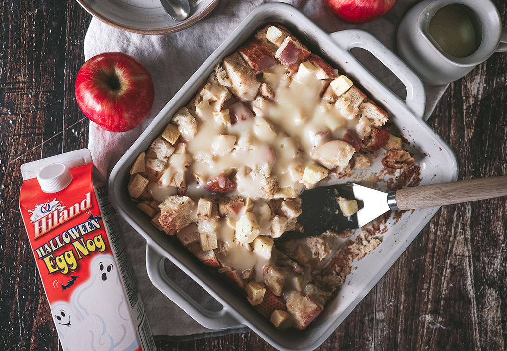 Baked apple bread pudding topped with eggnog sauce in a dish, next to a carton of Hiland Halloween Egg Nog and fresh apples.