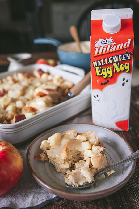 A plate of apple bread pudding served next to a carton of Hiland Halloween Egg Nog and a casserole dish of the dessert.