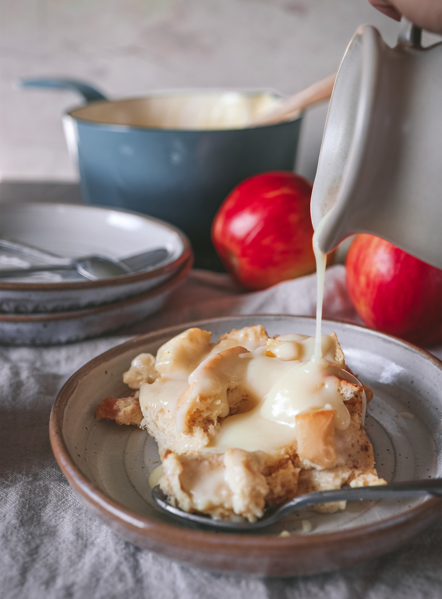 Close-up of creamy eggnog sauce being poured over apple bread pudding on a plate, with apples and a pot in the background.