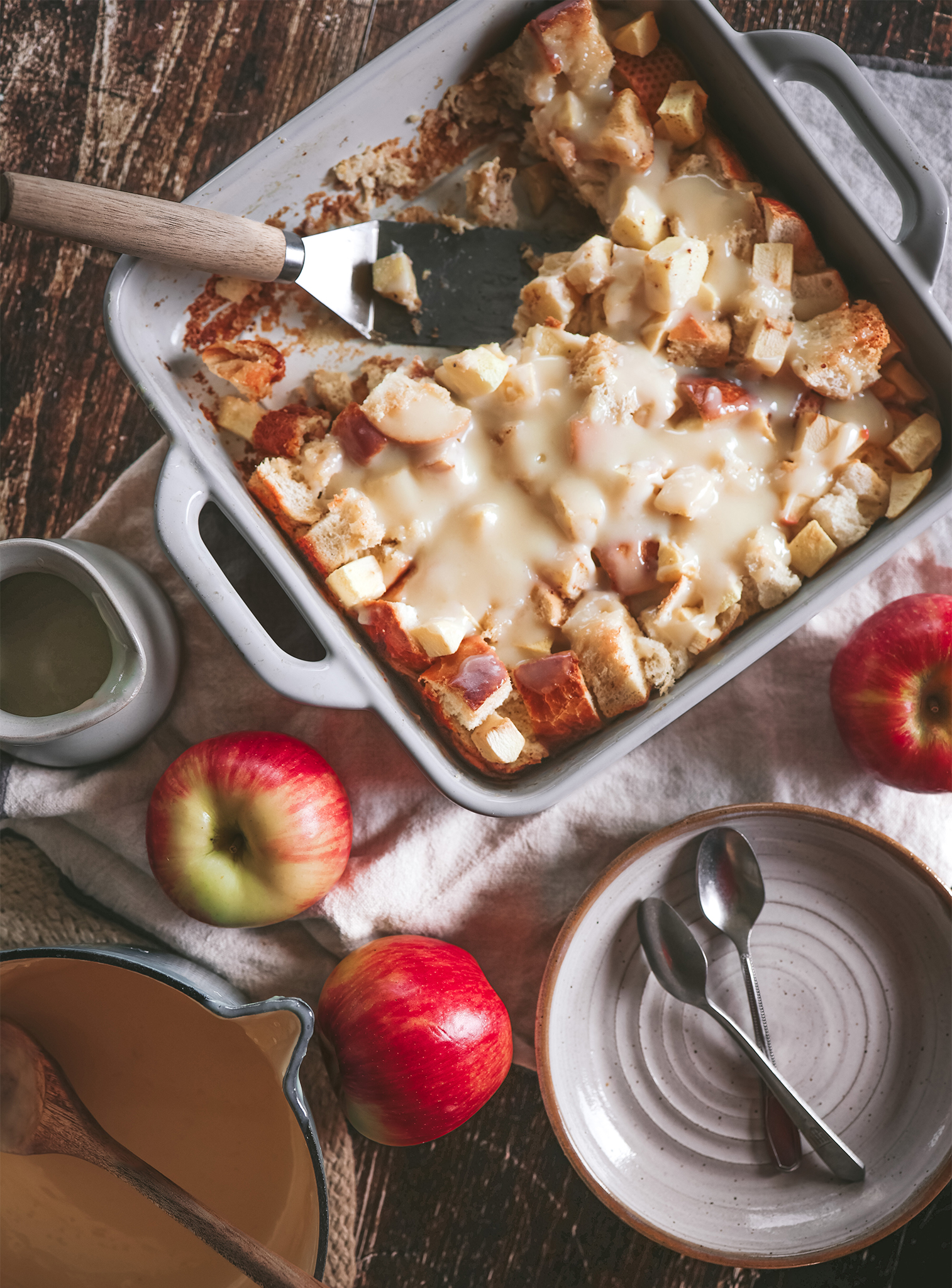 Baked apple bread pudding in a casserole dish with a serving spatula, surrounded by apples, bowls, and a jug of eggnog sauce.