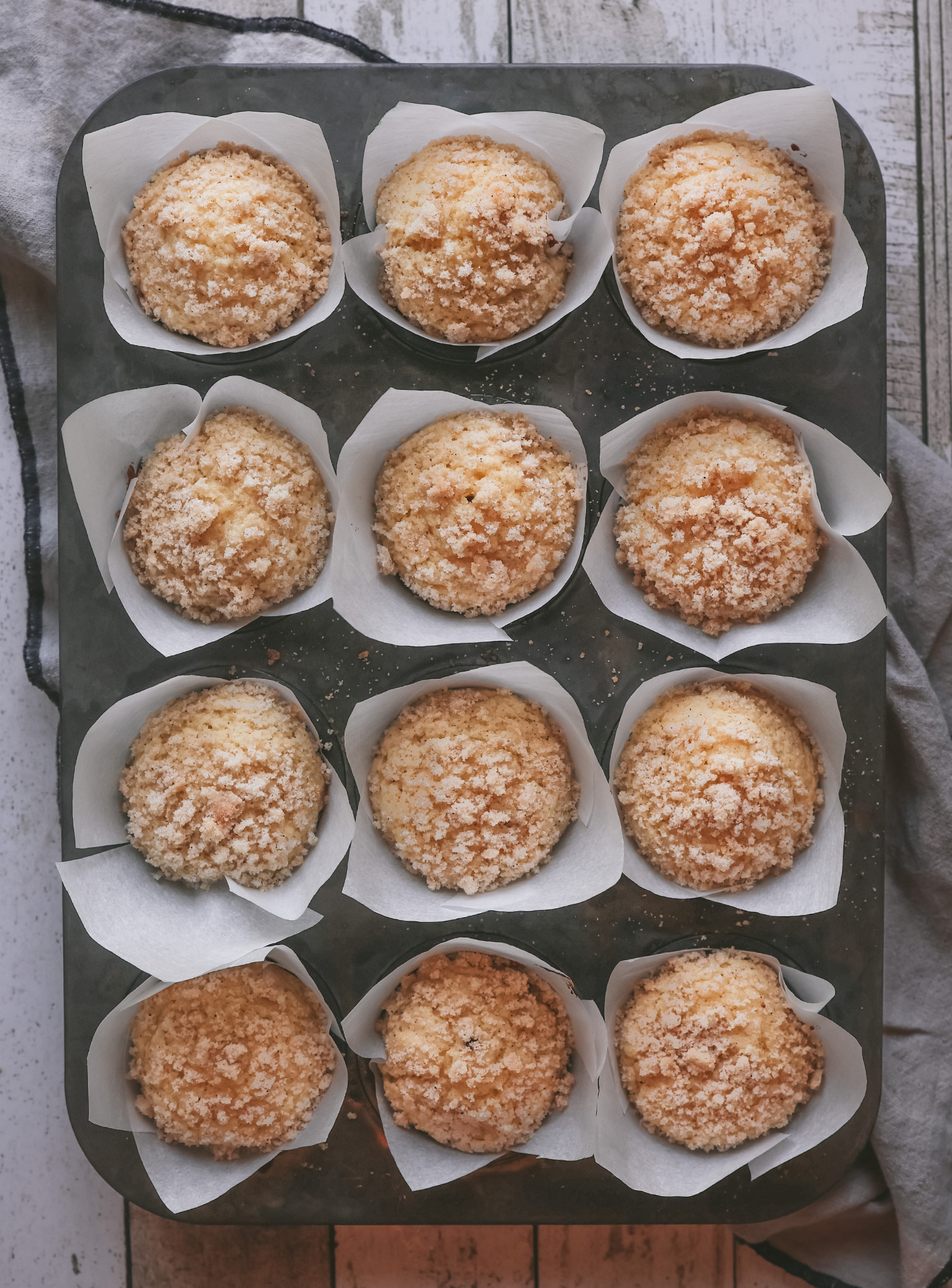 A muffin tin filled with twelve freshly baked eggnog muffins, each topped with a generous layer of sweet, crumbly streusel. The muffins are golden brown and nestled in white parchment paper liners.