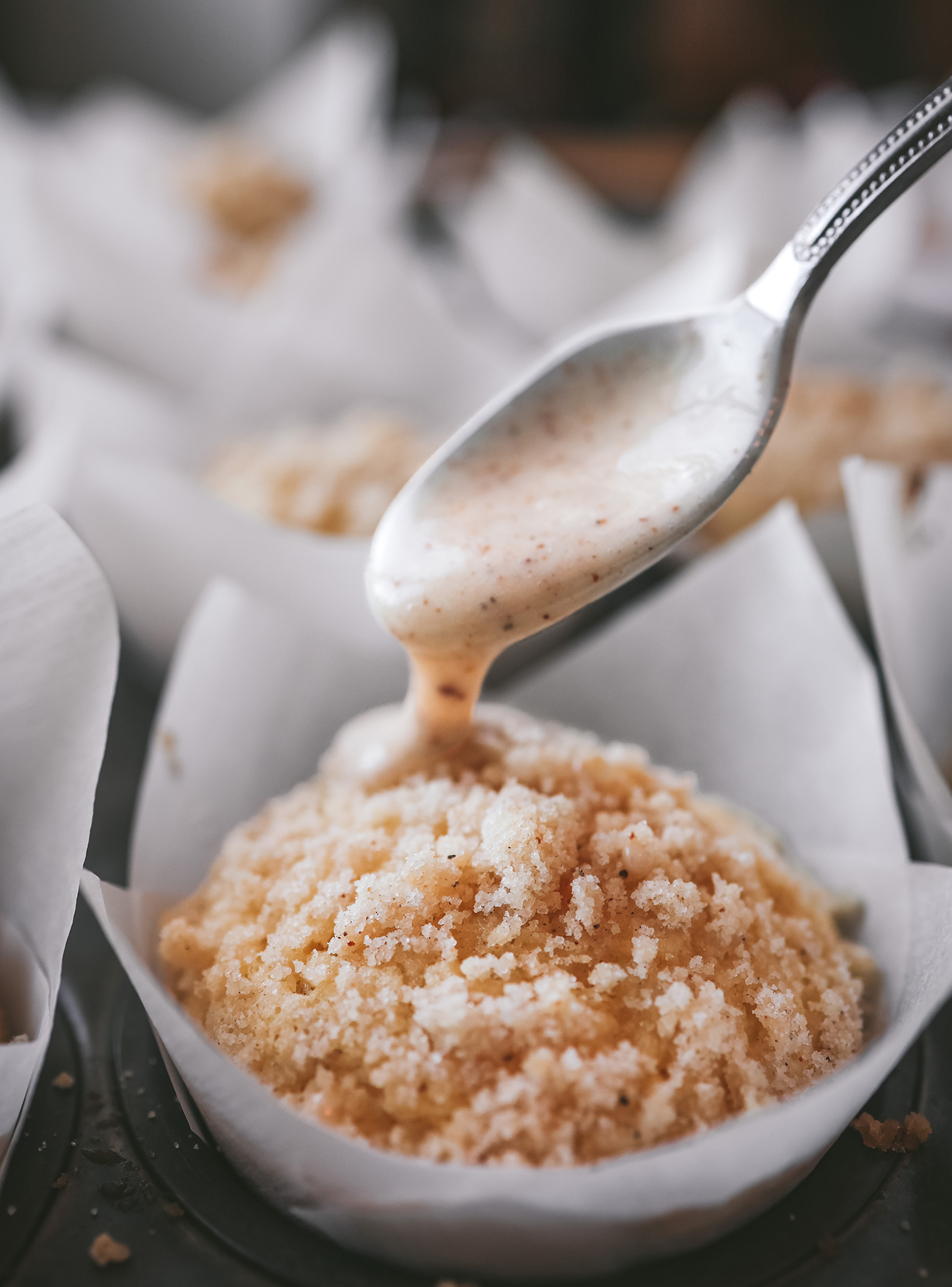 A close-up of a spoon drizzling creamy, eggnog glaze over an eggnog muffin with a crumbly streusel topping. The muffin rests in a parchment paper liner inside a dark muffin pan.