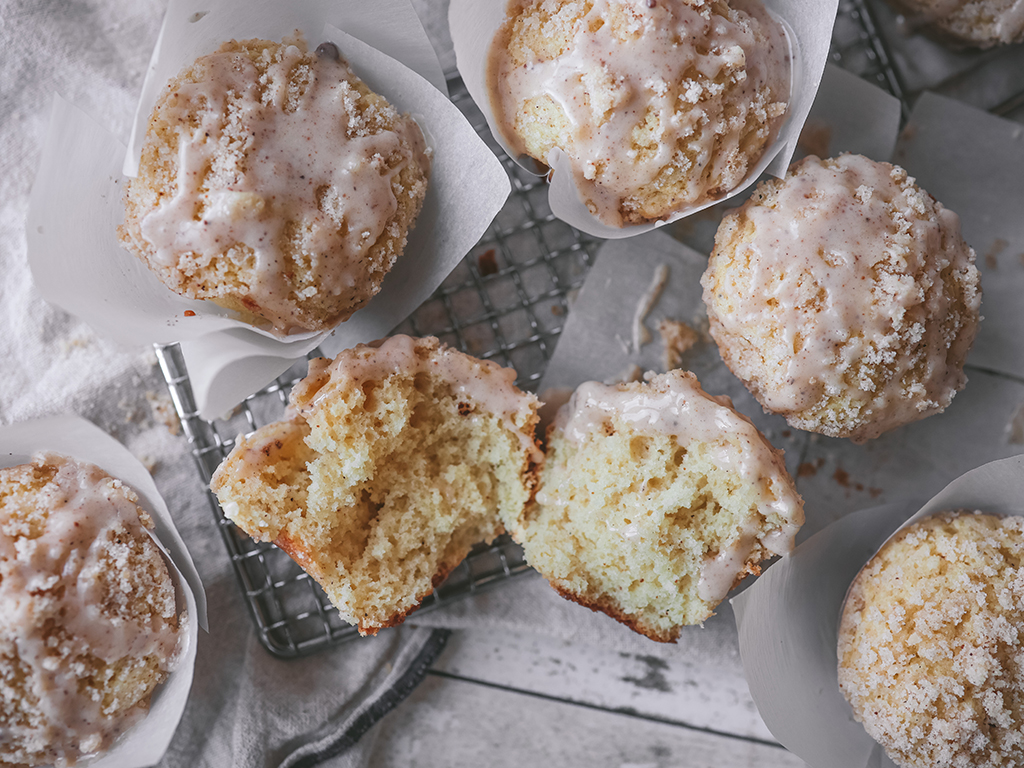 A close-up of a batch of eggnog muffins on a wire cooling rack, with one muffin torn in half to reveal its soft, fluffy interior. The muffins are topped with a sugary crumble and drizzled with a light eggnog and nutmeg glaze.