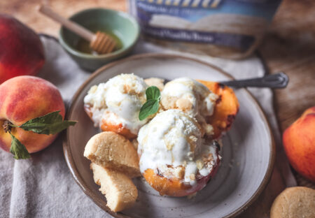 Grilled peach halves topped with scoops of Hiland Vanilla ice cream, sprinkled with cookie crumbles and drizzled with honey, served on a ceramic plate with shortbread cookies and a fresh mint garnish.