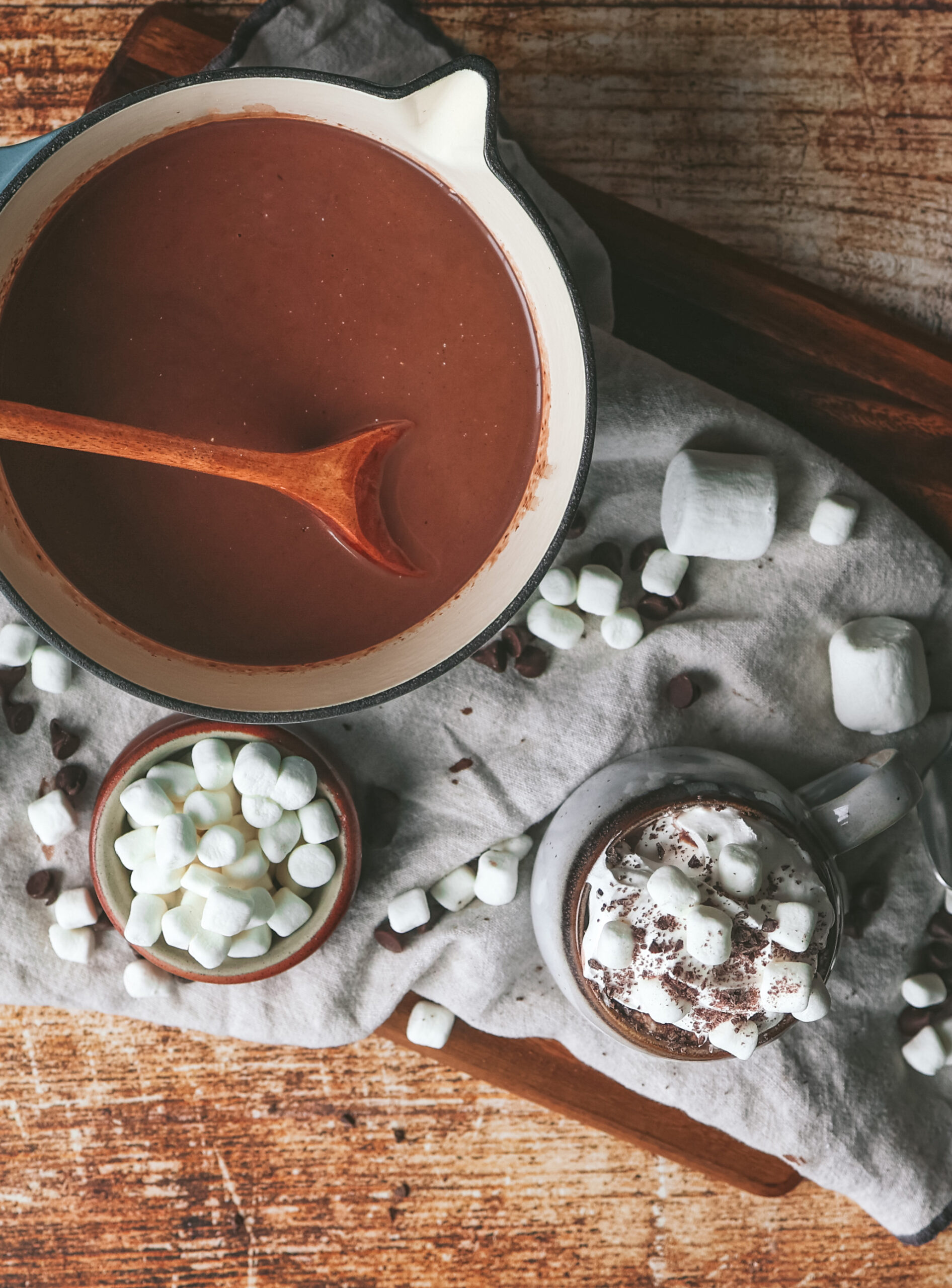 A cream-colored pot filled with rich, steaming hot chocolate sits on a rustic wooden surface, with a wooden spoon submerged in the drink. Surrounding the pot are mini marshmallows in a small bowl, scattered marshmallows, and a mug topped with whipped cream and chocolate shavings.