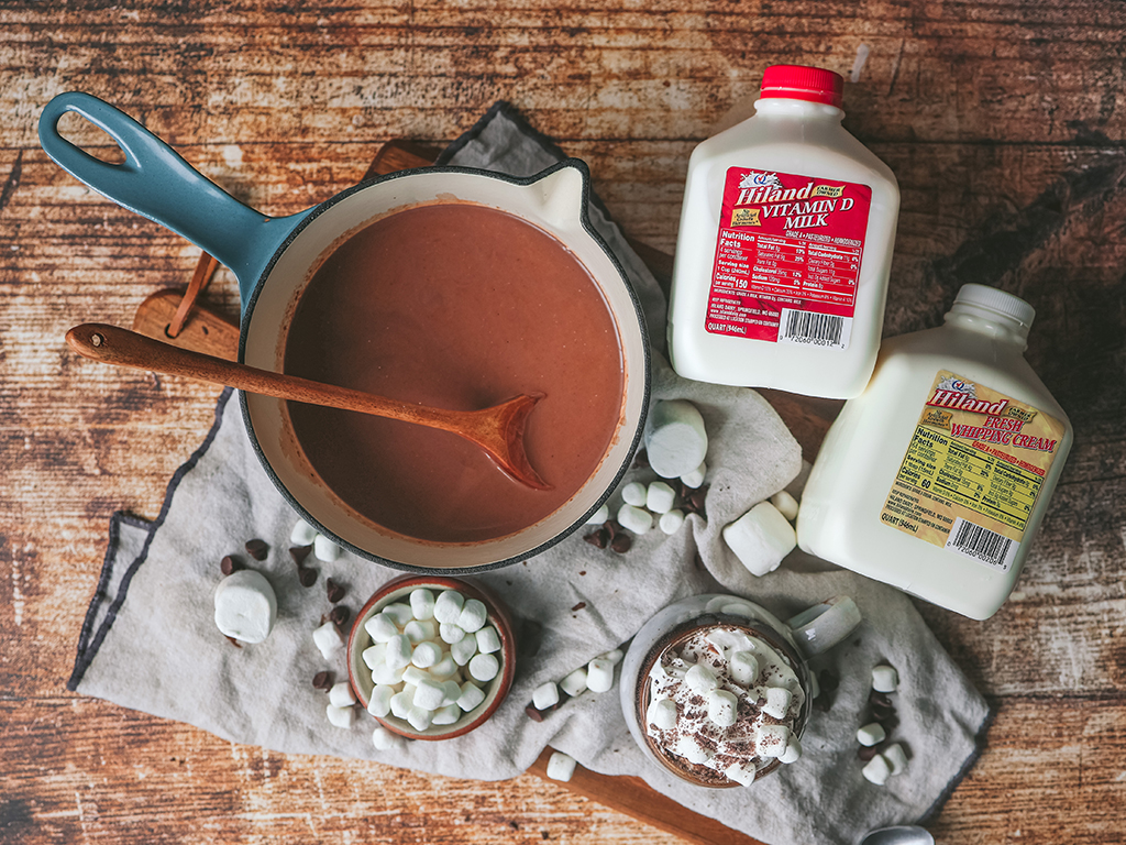 A top-down view of a pot filled with hot chocolate next to Hiland Dairy Vitamin D milk and fresh whipping cream cartons. Mini marshmallows, chocolate chips, and a whipped cream-topped mug complete the inviting setup on a light cloth backdrop.