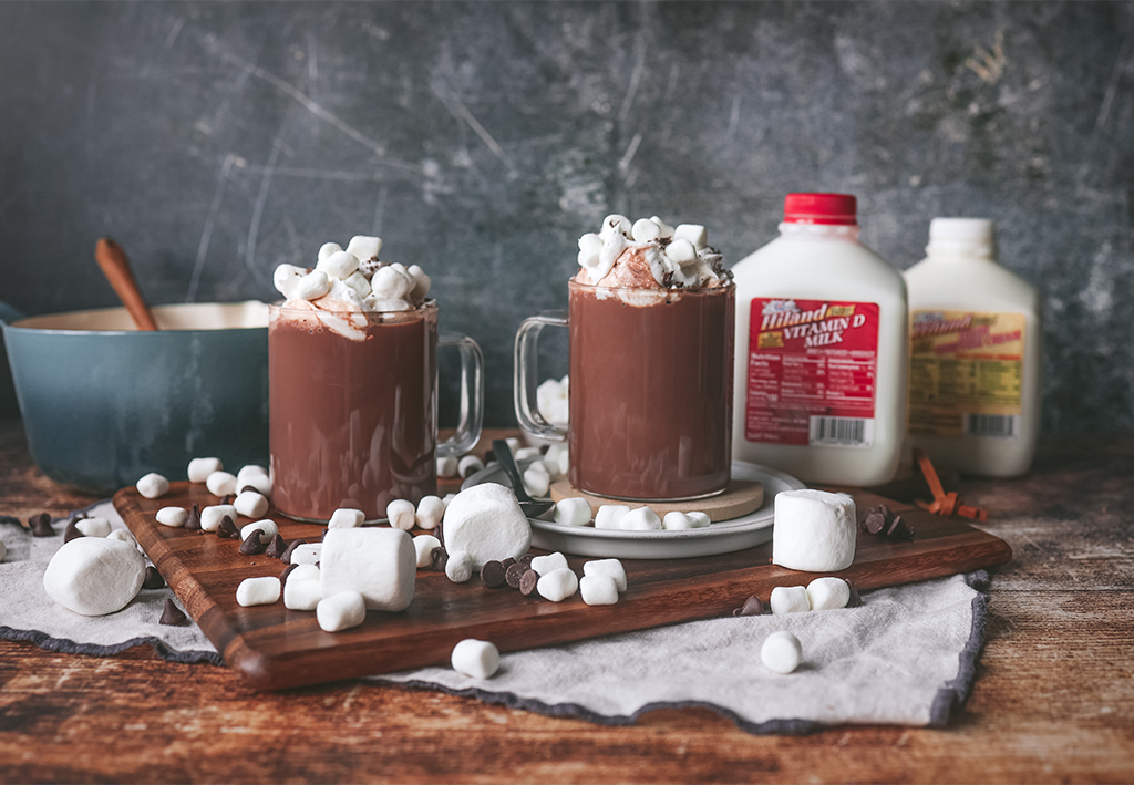 Two glass mugs filled with hot chocolate and topped with marshmallows sit on a wooden board surrounded by large marshmallows and chocolate chips. In the background are Hiland Dairy Vitamin D milk containers and a pot with hot chocolate, evoking a cozy winter scene.