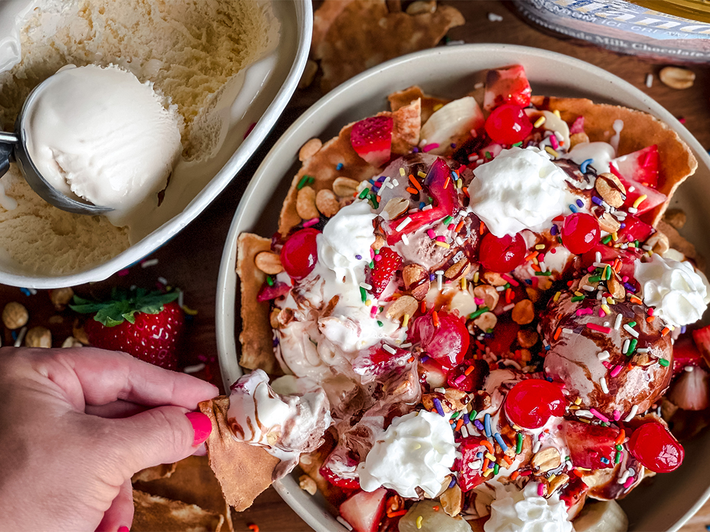 A plate of colorful ice cream nachos topped with scoops of vanilla and chocolate ice cream, whipped cream, cherries, sprinkles, strawberries, and nuts, with a hand holding a chip and an ice cream tub nearby.