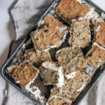 A metal baking tray filled with homemade ice cream sandwiches made from thick chocolate chip cookies and creamy vanilla ice cream. Some sandwiches are cut into squares, with slightly melted edges, creating a rustic, indulgent presentation. The tray rests on a soft linen cloth, adding to the homemade feel.