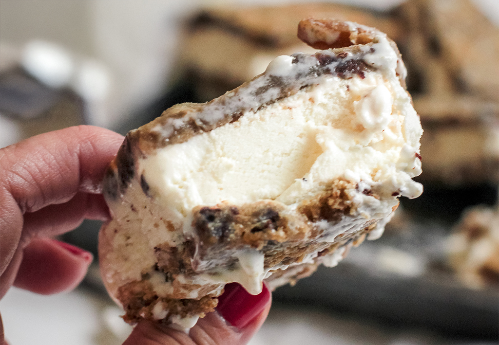 A close-up of a hand holding a homemade ice cream sandwich with thick, creamy vanilla ice cream between two chewy chocolate chip cookie layers. The ice cream is slightly melting, making the treat look even more indulgent.