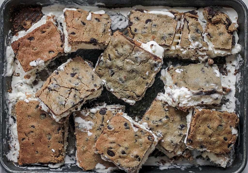 A baking tray filled with homemade ice cream sandwiches made from thick chocolate chip cookie slabs and creamy vanilla ice cream. The sandwiches are cut into squares, with some pieces slightly melting, creating a deliciously messy look.