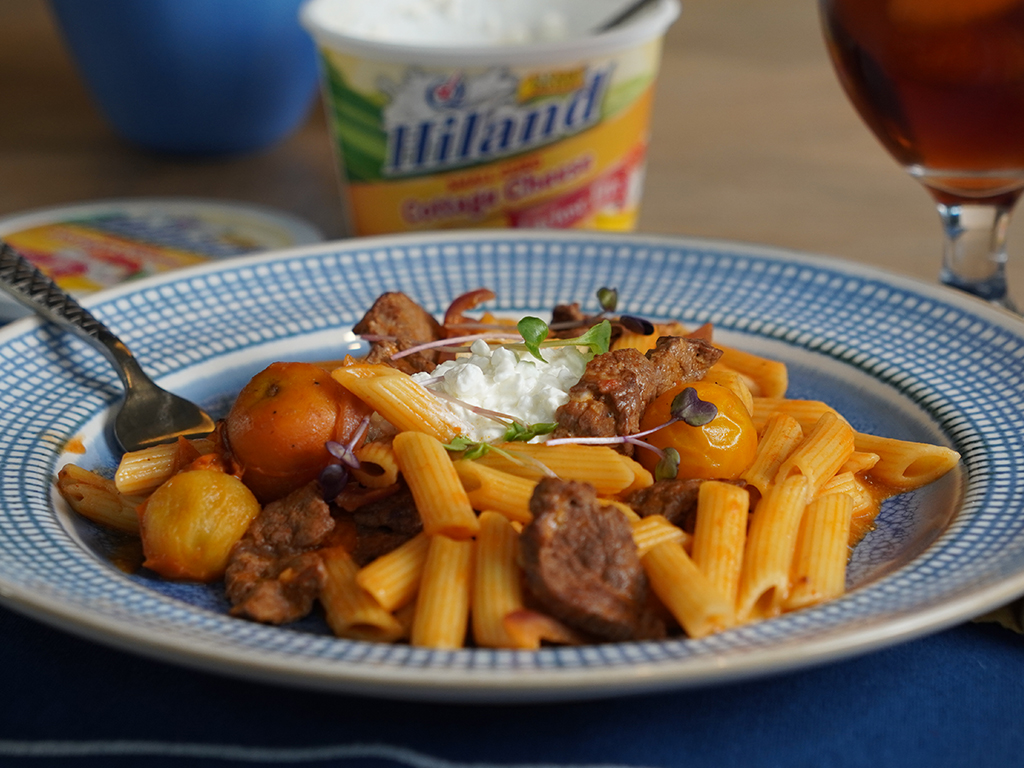 A plate of penne pasta with a sauce made with Hiland Dairy Lactose Free Cottage Cheese with tender beef, cherry tomatoes, garnished with microgreens, served on a blue and white patterned plate.