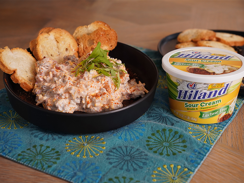 Creamy lactose-free dip made with Hiland Lactose Free Sour Cream, topped with paprika and garnished with microgreens, served with toasted bread slices in a black bowl.