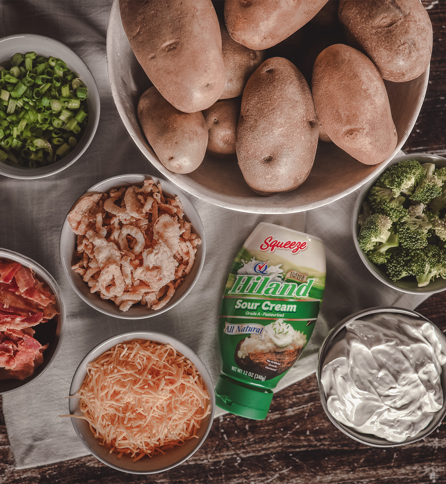 A bowl of raw potatoes surrounded by small bowls of mashed potato toppings, including green onions, crispy onions, shredded cheese, broccoli, bacon, and sour cream, with a bottle of Hiland Sour Cream at the center.