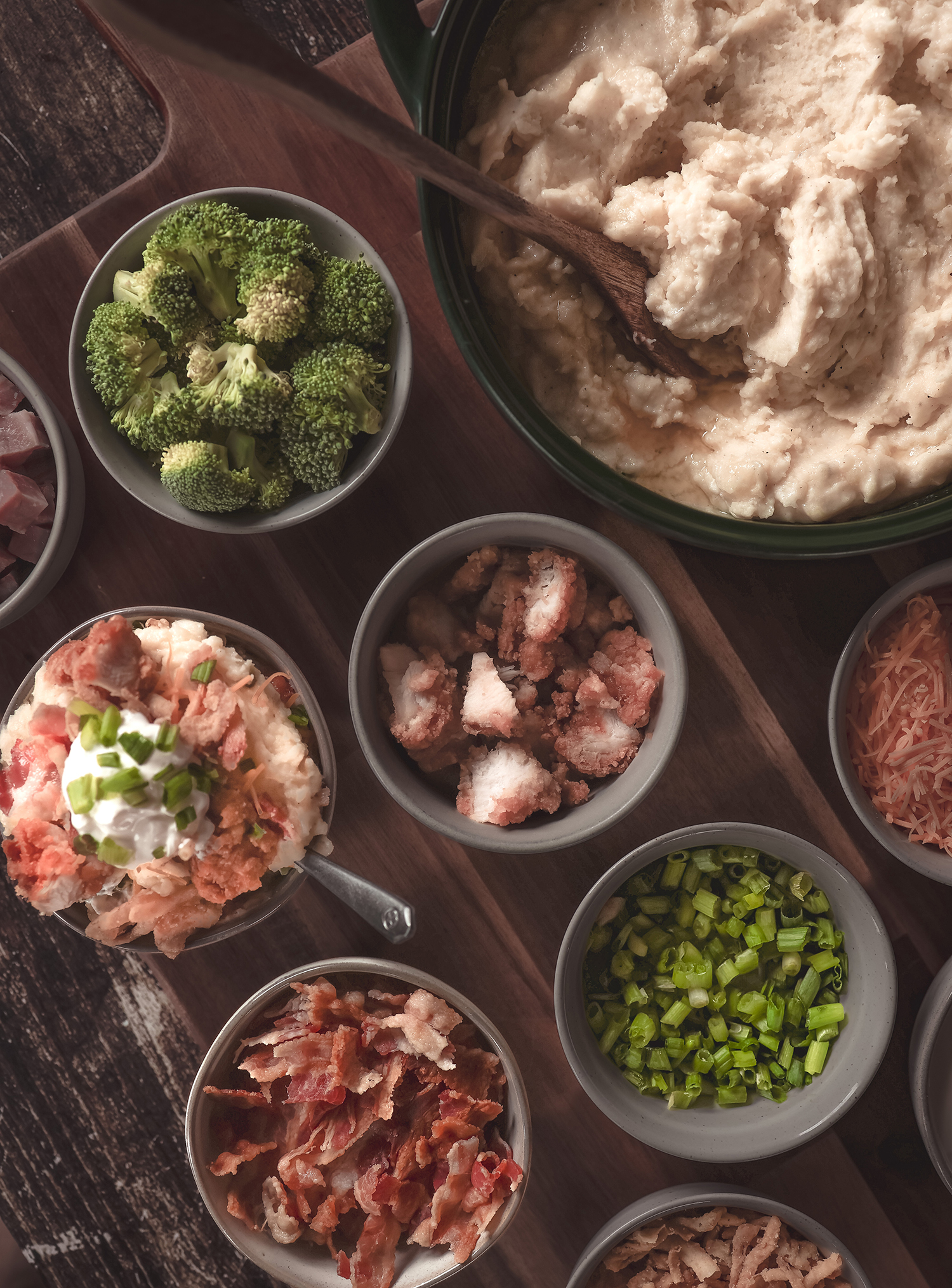 An overhead view of a mashed potato bar setup with bowls of toppings like bacon, broccoli, green onions, shredded cheese, and ham, alongside a pot of mashed potatoes.