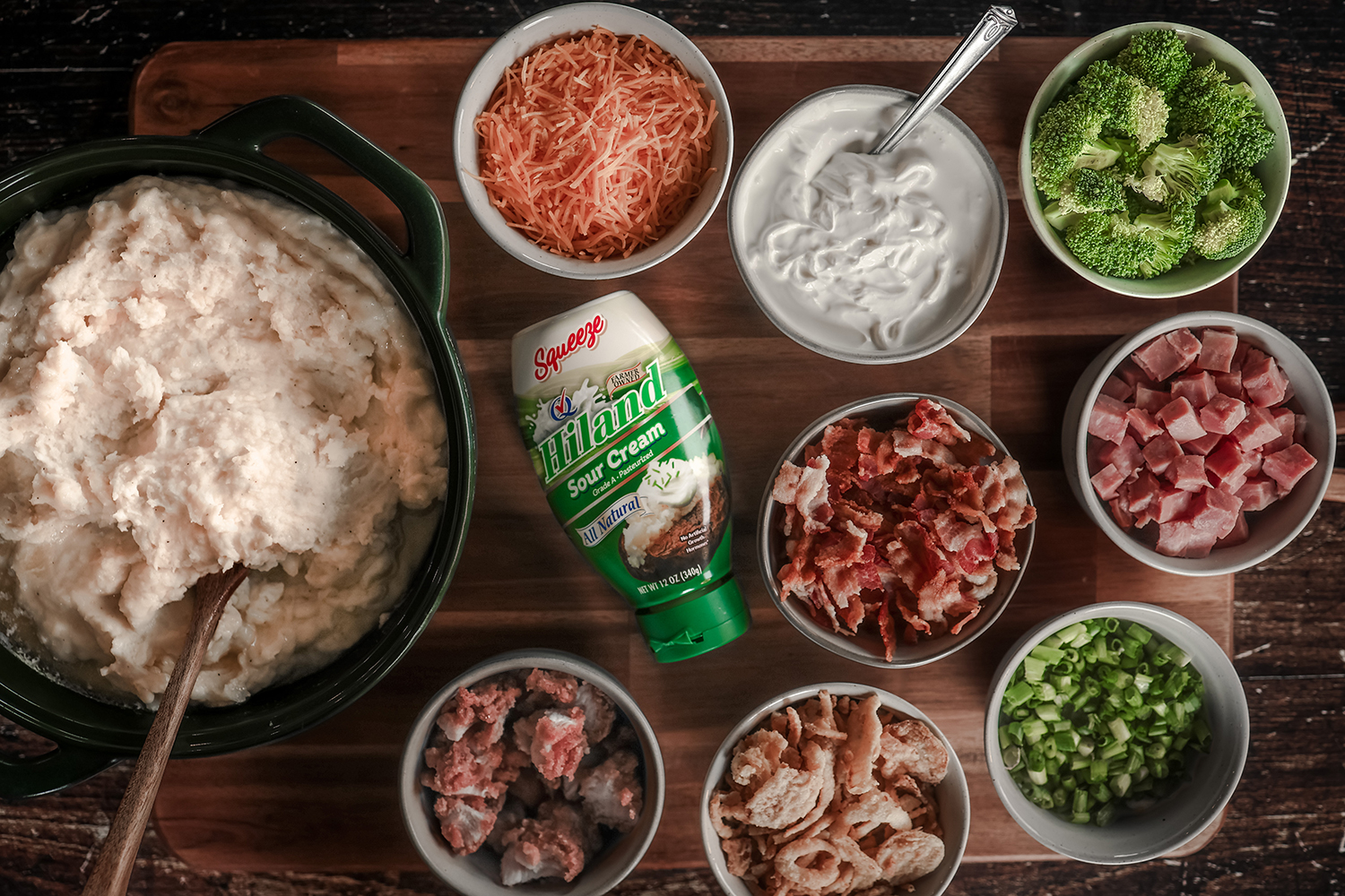 An overhead view of a mashed potato bar featuring Hiland Sour Cream, a pot of mashed potatoes, and bowls of assorted toppings such as broccoli, shredded cheese, diced ham, bacon, green onions, and crispy onions.