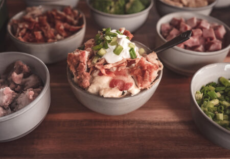 A close-up of a bowl of mashed potatoes with various toppings like bacon, green onions, sour cream, and crispy onions, surrounded by other topping bowls.