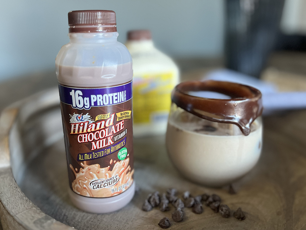 A bottle of Hiland Chocolate Milk with a rich, creamy appearance sits on a rustic wooden tray. Beside it, a glass filled with chocolate milk has a thick layer of chocolate coating dripping from the rim. A small pile of chocolate chips is scattered in the foreground, adding to the indulgent feel of the scene. In the slightly blurred background, another dairy product container and a folded cloth are visible, enhancing the cozy and inviting atmosphere.