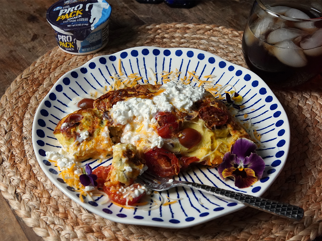 Protein vegetable omelette topped with cottage cheese and shredded cheese, served with roasted cherry tomatoes on a blue and white patterned plate, with a container of Pro Pack Cottage Cheese and a glass of iced beverage in the background.