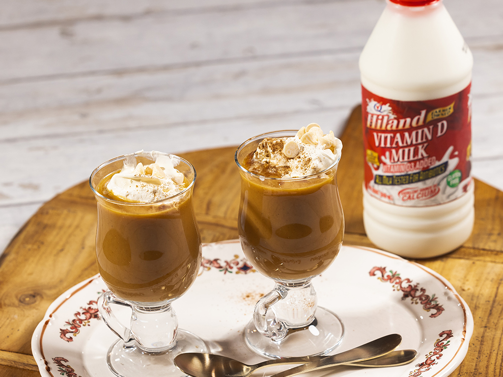 Two glasses of pumpkin spice white hot chocolate topped with whipped cream, served on a decorative plate with Hiland Vitamin D Milk in the background.