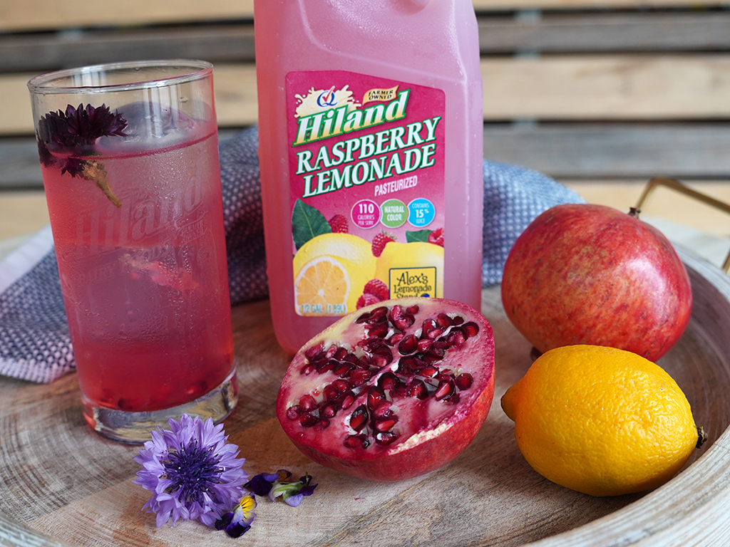 A chilled glass of raspberry lemonade cocktail garnished with a flower sits beside a bottle of Hiland Raspberry Lemonade, surrounded by a halved pomegranate, whole pomegranate, lemon, and a few fresh blossoms on a wooden tray.