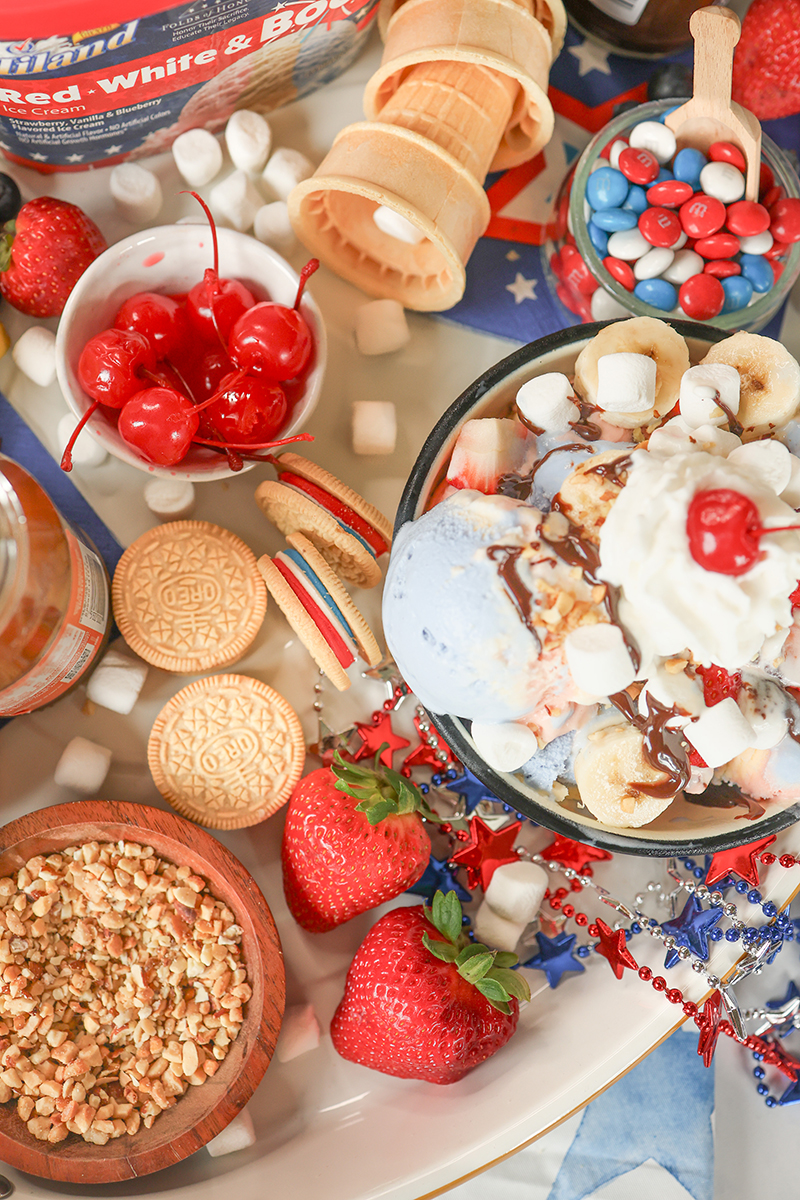 Overhead view of a patriotic ice cream sundae board featuring Hiland Red, White & Boom Ice Cream with bananas, strawberries, cherries, marshmallows, cookies, chopped nuts, chocolate sauce, candy-coated chocolates, and festive red, white, and blue decorations.