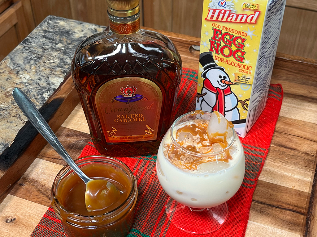 A glass of eggnog drizzled with caramel sauce sits beside a jar of caramel, a bottle of salted caramel whiskey, and a carton of Hiland eggnog.