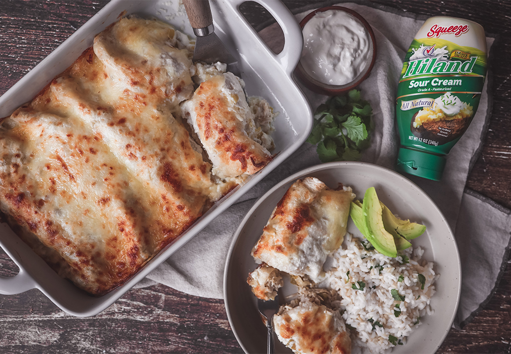 Sour cream chicken enchiladas in a baking dish, served with a side plate featuring rice, avocado slices, and a portion of the enchiladas. A bottle of Hiland Sour Cream and fresh cilantro are nearby.