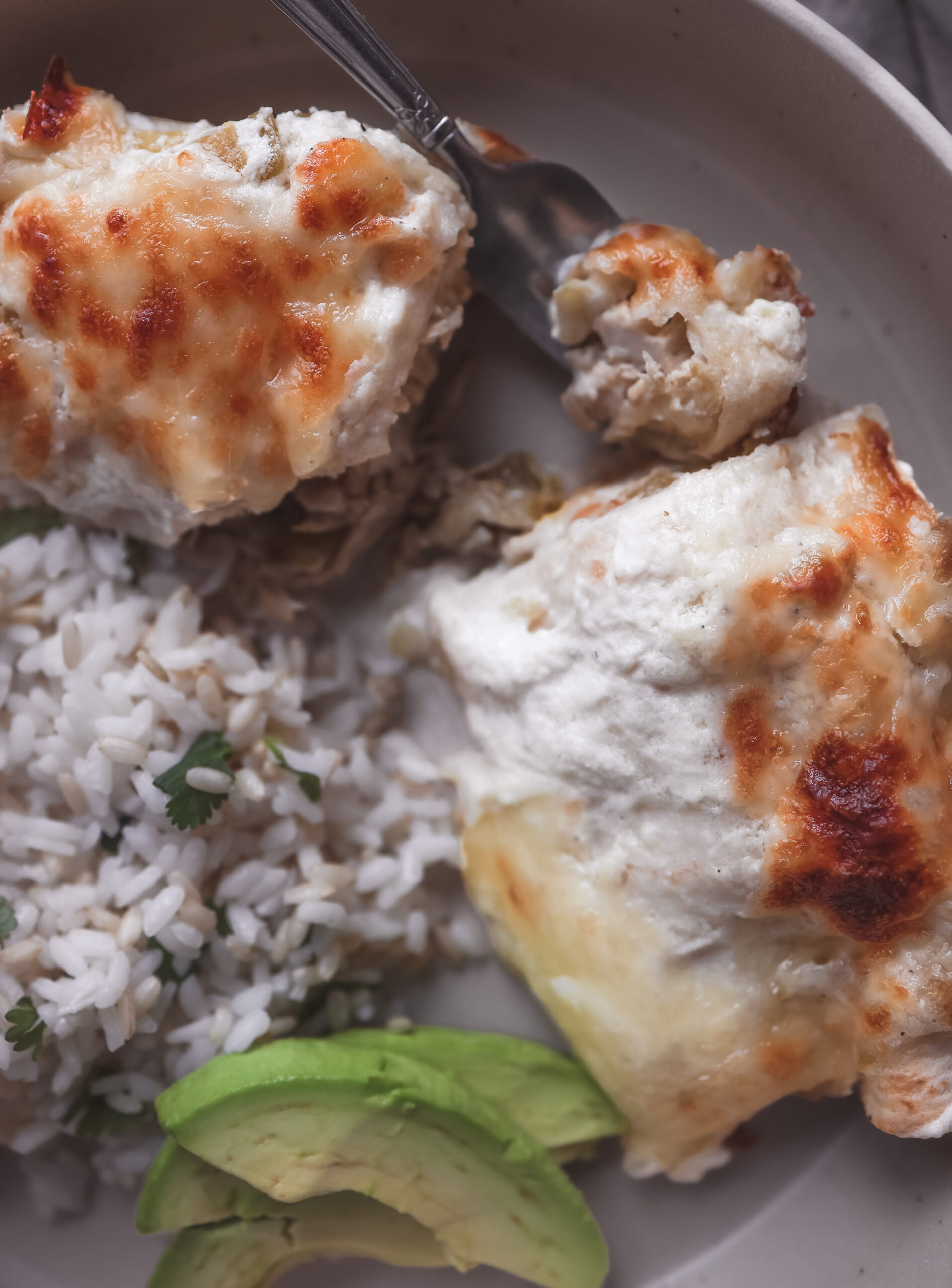 A close-up of a plate with two sour cream chicken enchiladas served with white rice and fresh avocado slices. The enchiladas are topped with melted, golden-brown cheese.