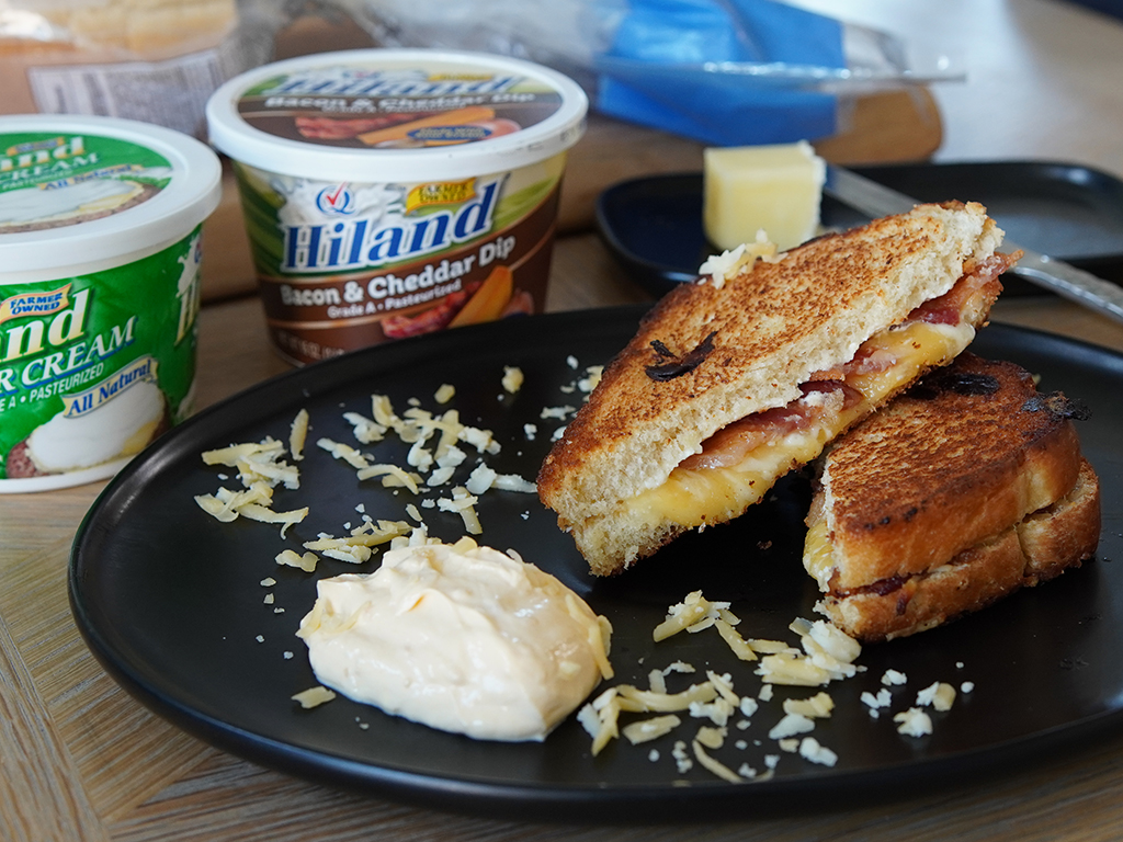 Grilled cheese sandwich with bacon and cheddar, served on a black plate with shredded cheese and a dollop of dip, alongside Hiland sour cream and bacon & cheddar dip containers.