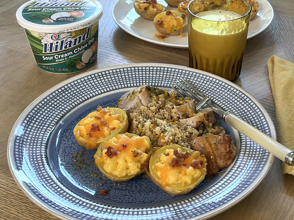 A plate featuring twice-baked potatoes topped with melted cheese and bacon, served with a side of pork and stuffing, next to a Hiland sour cream chive dip container and a glass of milk.