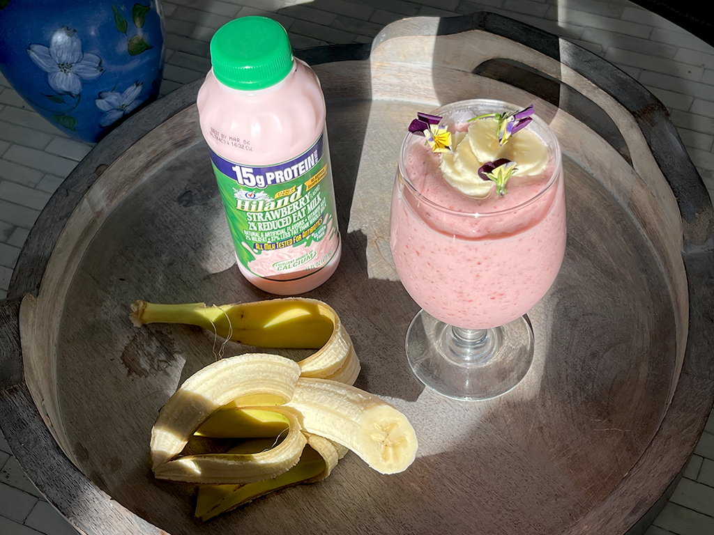 A rustic wooden tray holds a refreshing strawberry banana smoothie in a stemmed glass, garnished with banana slices and delicate edible flowers. Next to it, a bottle of Hiland Strawberry 2% Reduced Fat Milk sits in the sunlight, emphasizing its creamy pink hue. A partially peeled banana rests beside the smoothie, adding a natural element to the composition. The bright lighting and soft shadows create a warm and inviting scene.