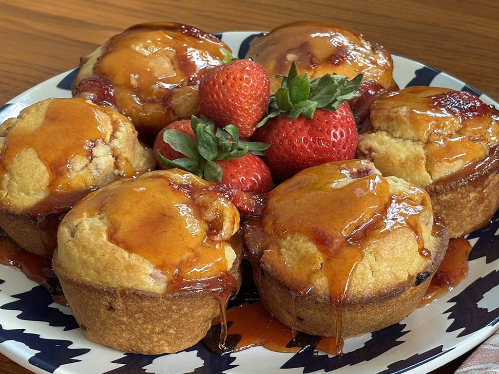 Strawberry cornbread muffins drizzled with a glossy strawberry glaze and arranged on a platter around fresh strawberries, with extra glaze pooled around the muffins.