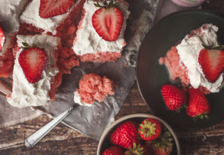 Overhead view of sliced strawberry tres leches cake served on parchment. The cake is topped with whipped cream and halved fresh strawberries. A fork with a bite of cake rests on the tray, and a small plate of cake with fresh strawberries sits nearby on a rustic wood table.