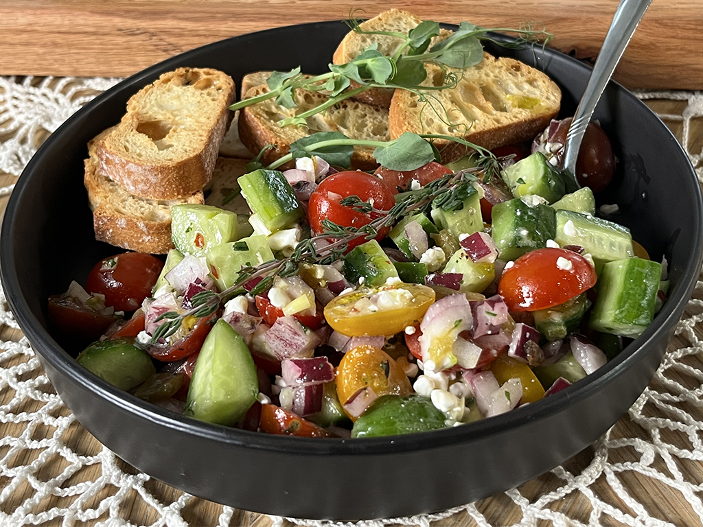 Fresh tomato cucumber salad with cherry tomatoes, diced cucumbers, red onion, and crumbled cottage cheese, garnished with thyme and served with toasted baguette slices in a black bowl.