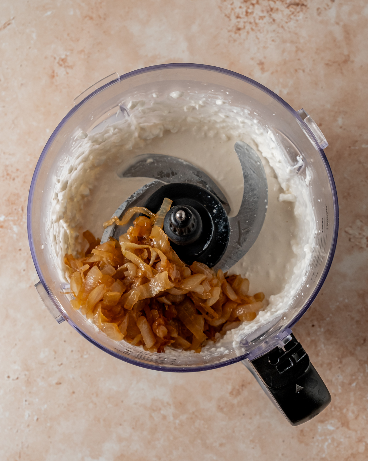 Food processor with whipped mixture of cottage cheese and sour cream, with caramelized onions on top, ready to be blended.