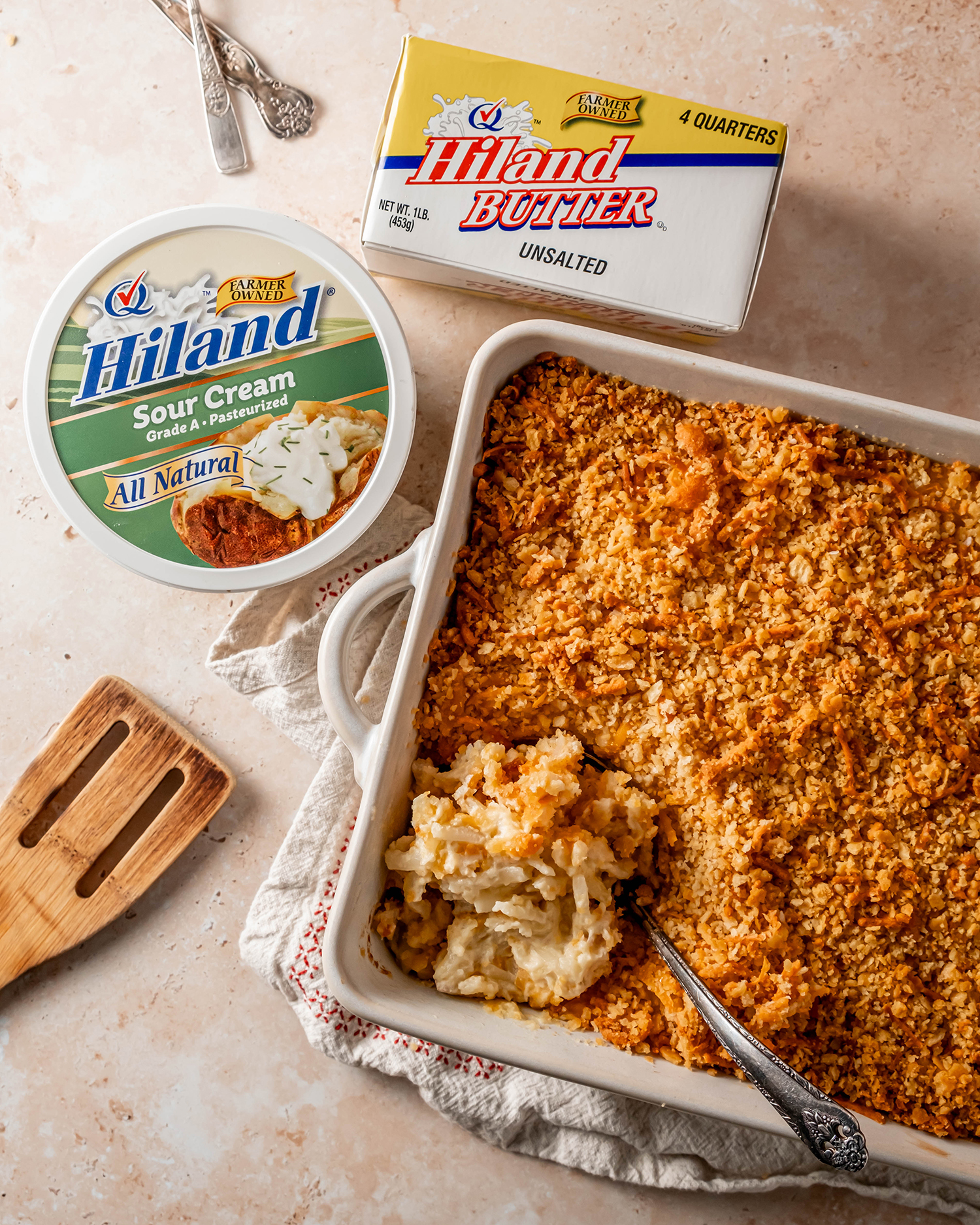 Cheesy potato casserole served in a white dish, displayed alongside with Hiland sour cream and butter a wooden spatula and cutlery.