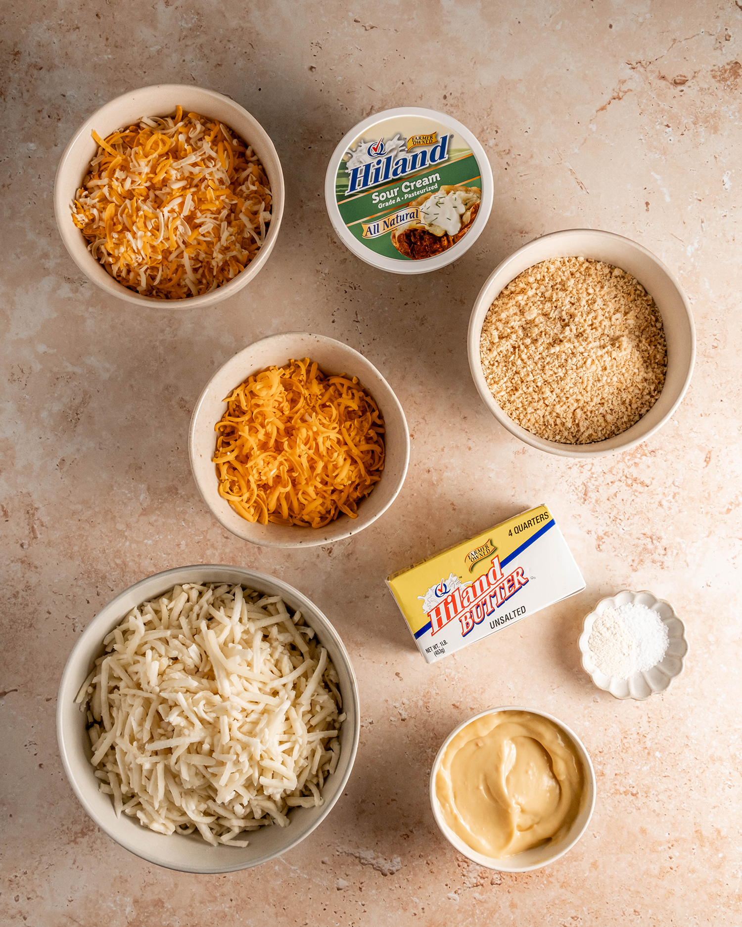 Ingredients for a cheesy potato casserole laid out, including shredded potatoes, cheese, sour cream, butter, and breadcrumbs.