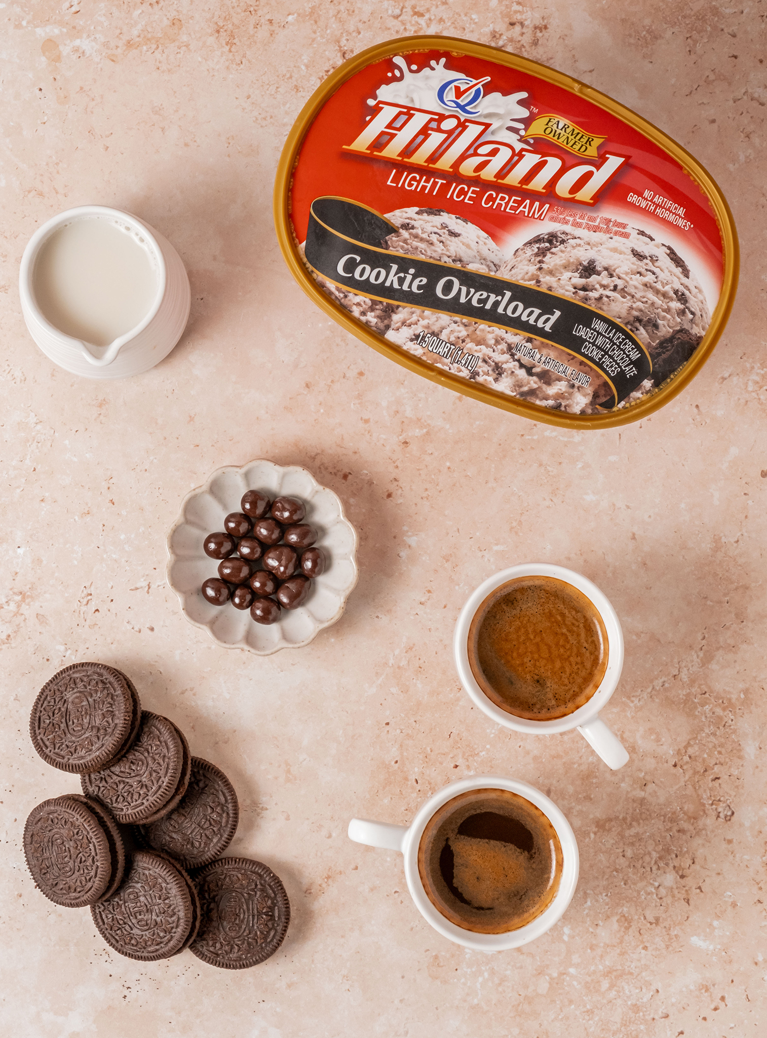 A flat lay of ingredients on a tan stone surface including a container of Hiland Cookie Overload Light Ice Cream, chocolate sandwich cookies, chocolate-covered espresso beans, espresso shots, and a small pitcher of milk.